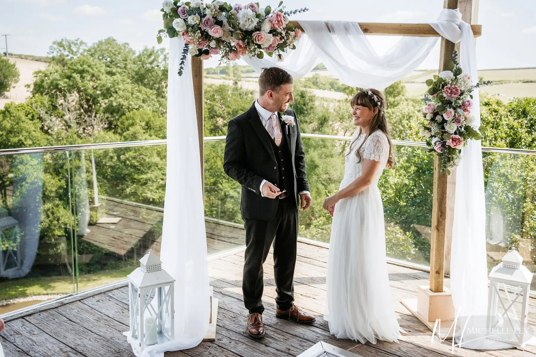 A couple smiling at each other under a floral arch at Tree Top Escape budget wedding venue in Devon