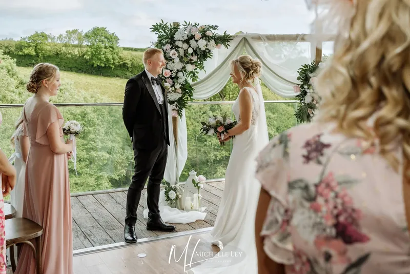 Couple exchanging vows under a floral arch in The Nest, with views over the North Devon countryside